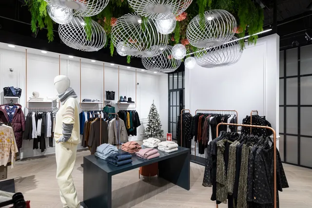 Interior of a modern apparel store with garment rails, folded clothing displays and a fitting-room corridor under overhead ceiling cameras